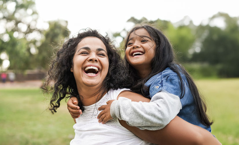a mother giving her daughter a piggyback ride. they both laugh. it is summer and there is greenery all around
