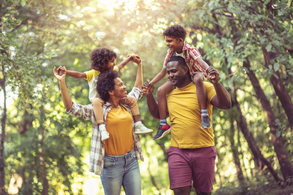 A joyful family walking through a sunlit forest. The parents each carry a child on their shoulders, smiling and holding hands as they enjoy the outdoors together, symbolizing unity and happiness.