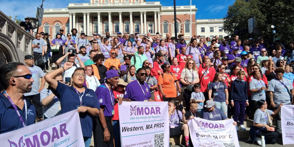 A large outdoor gathering of people wearing colorful shirts—purple, red, blue, gray, and orange—assembled on the steps of a historic building. They hold banners for MOAR (Massachusetts Organization for Addiction Recovery) that read “Join the Voices for Recovery,” symbolizing unity and advocacy for addiction recovery awareness.