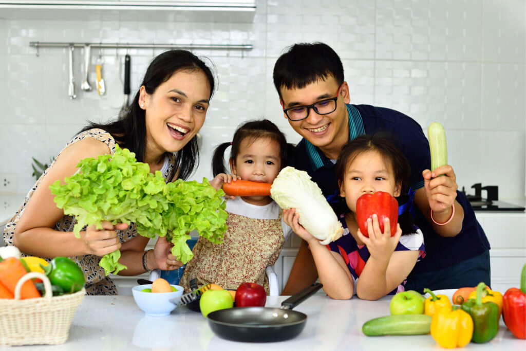 A smiling family in a kitchen holding fresh vegetables, surrounded by colorful produce on the counter.